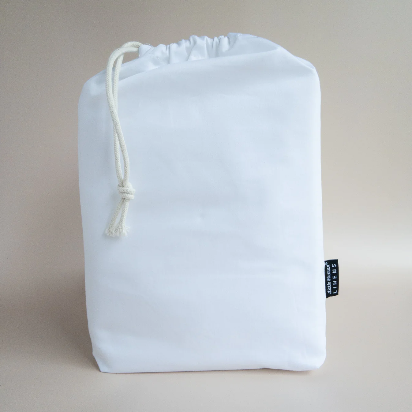 A white drawstring bag with a thick white cord, standing upright against a neutral beige background—perfect for storing your Little Human Linens waterproof sheet after use. A small black tag is attached to the side of the bag.