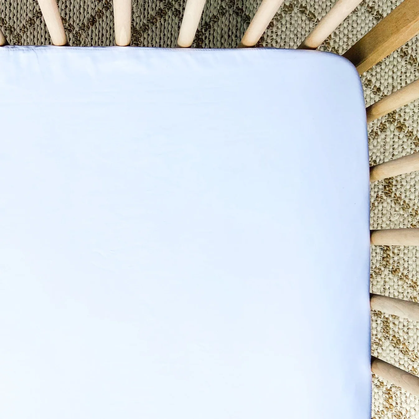 A close-up of a white crib mattress with wooden crib slats and a woven beige rug visible in the background, perfect for a Little Human Linens waterproof sheet review.