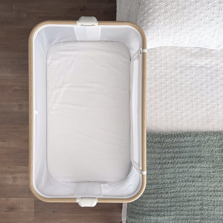 A top-down view of an empty baby bassinet with a white mattress, featuring a Little Human Linens waterproof sheet review, placed next to a bed with a textured white blanket and green knitted throw on a wooden floor.