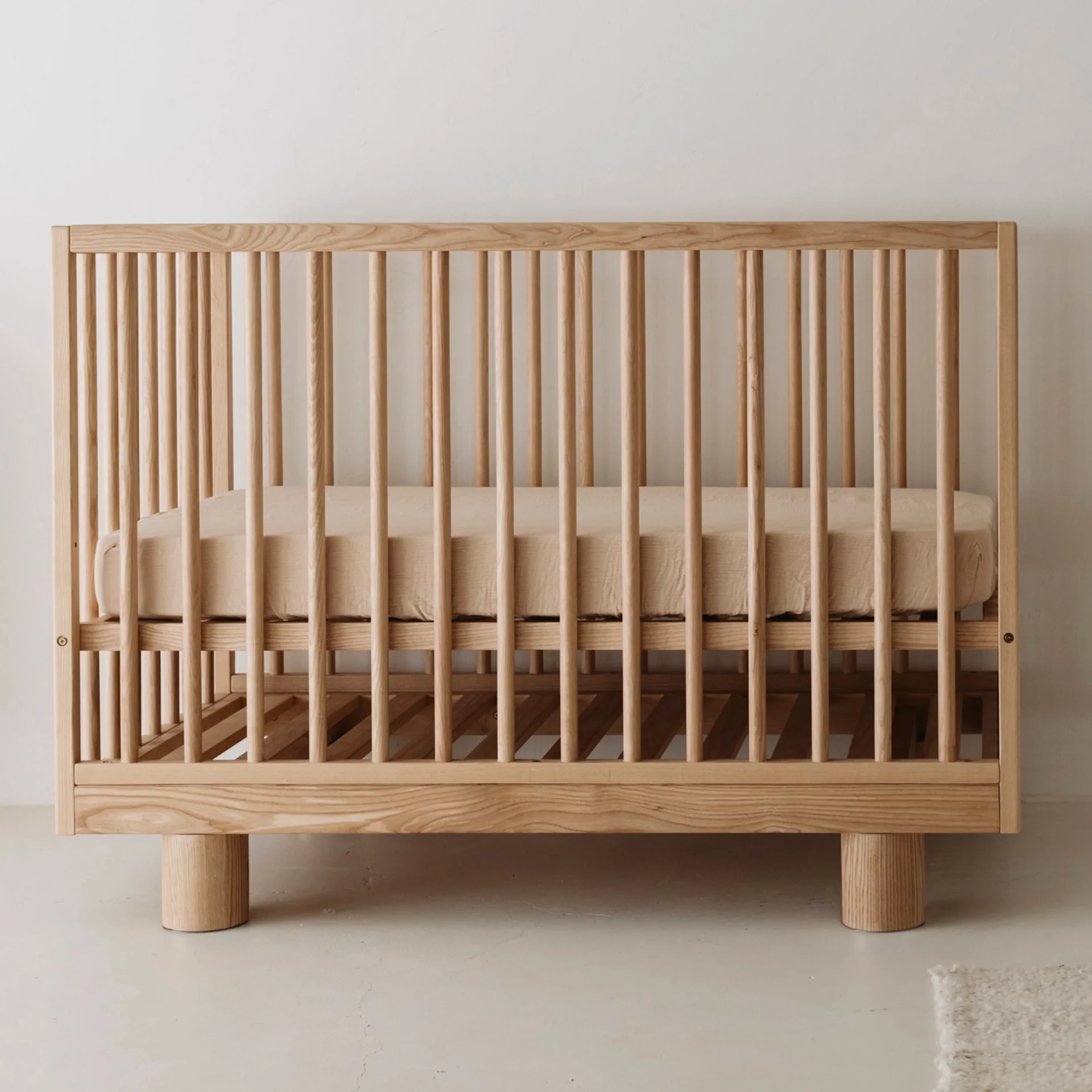 A minimalist wooden baby crib with vertical slats and a neutral-colored mattress, placed against a plain white wall on a light floor—perfect for those seeking simplicity inspired by the Hendrix Ashwood Cot review.