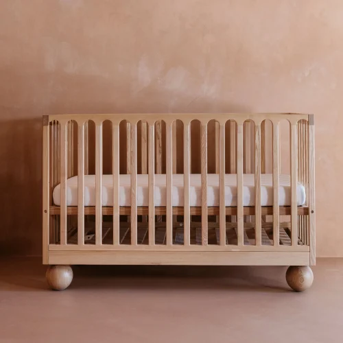 A Zali Ashwood Cot with vertical slats and large round legs sits against a beige textured wall and floor. This modern crib features a simple design and white mattress, perfect for any nursery. Read our Zali Ashwood Cot review for more details.