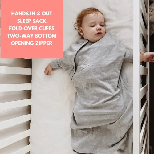 A baby sleeps in a gray Baby Loves Sleep Hands In & Out Sleep Sack inside a white crib. A pink box in the corner lists features: “Hands in & out sleep sack, fold-over cuffs, two-way bottom opening zipper.”.