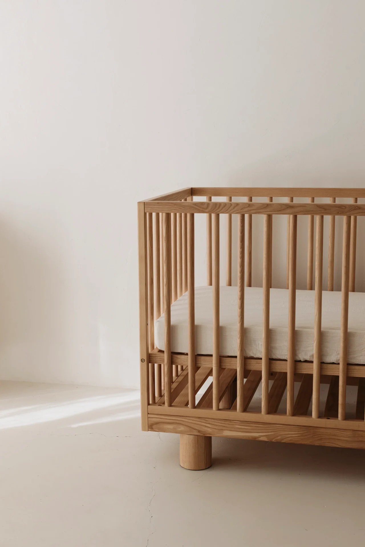 A minimalist wooden baby crib with vertical slats and a white mattress sits in a bright, empty room—perfect for those seeking a sleek nursery style, as noted in the latest Hendrix Ashwood Cot review.