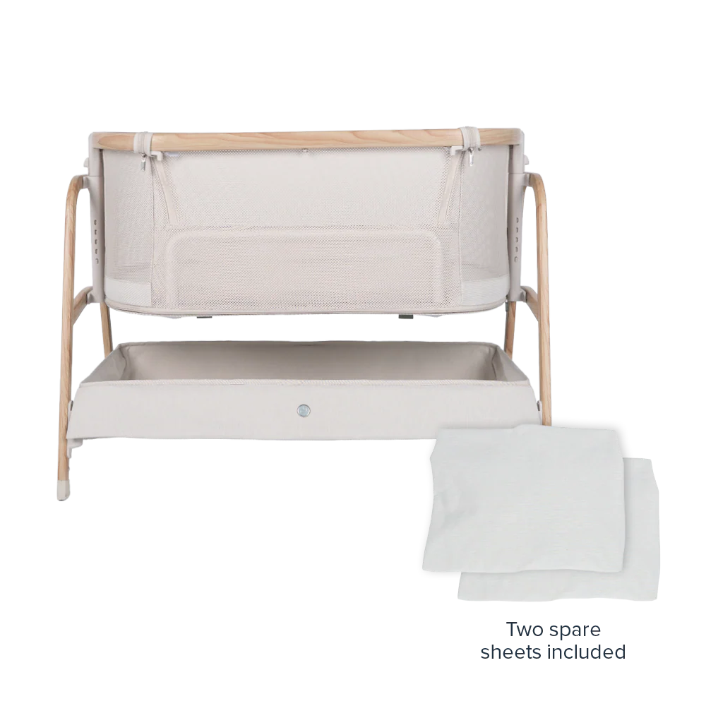 A modern Babybee Pod Bedside Sleeper with a light wood frame, mesh sides, a storage shelf underneath, and two folded white sheets placed in front.