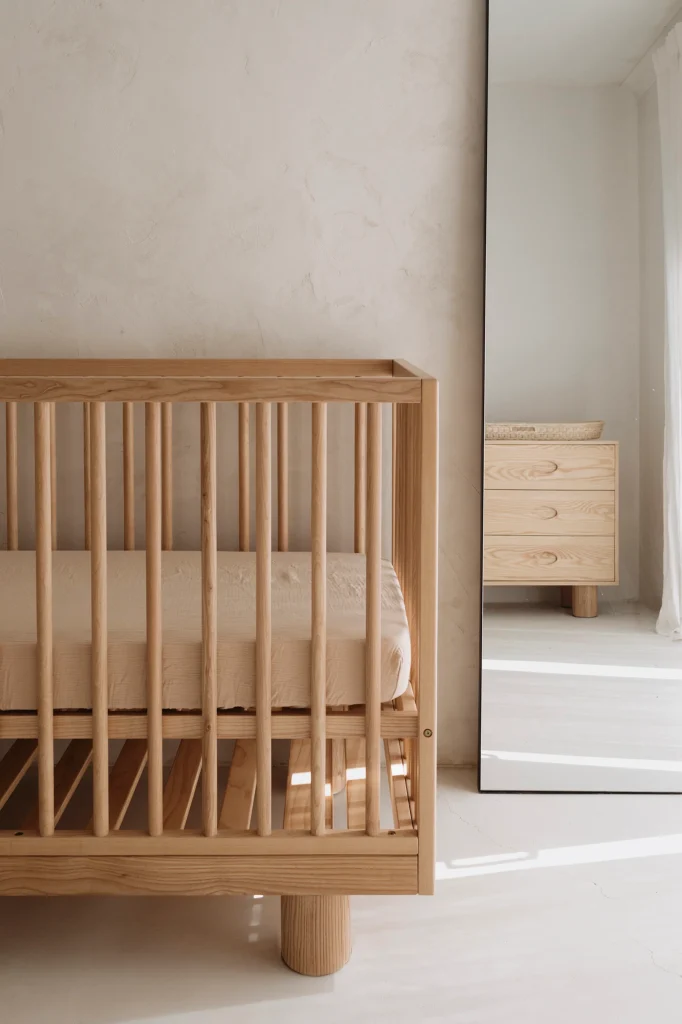A minimalist nursery with a wooden crib reminiscent of those praised in Hendrix Ashwood Cot reviews, a beige mattress, and a large mirror reflecting a matching wooden dresser. The room features light-colored walls and soft natural lighting.