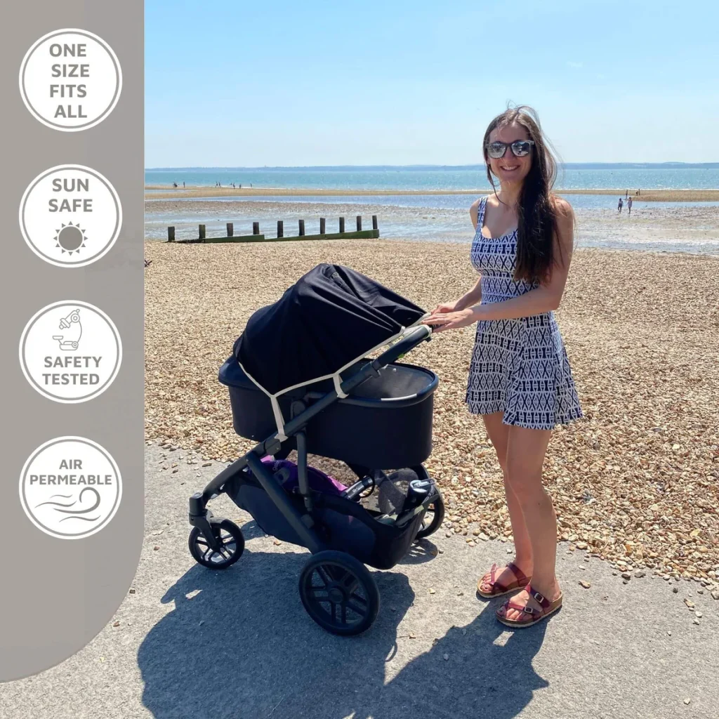 A woman in sunglasses stands on a pebbly beach, smiling next to a baby stroller with a sun cover. The sea is in the background. Icons highlight sun safety, safety-tested, and air permeable features—perfect for any SnoozeShade pram shade review.