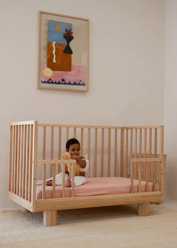 A young child sits in a Hendrix Ashwood Cot with a pink mattress, holding a teddy bear. The room features neutral tones, and a colorful abstract painting hangs on the wall above the crib.