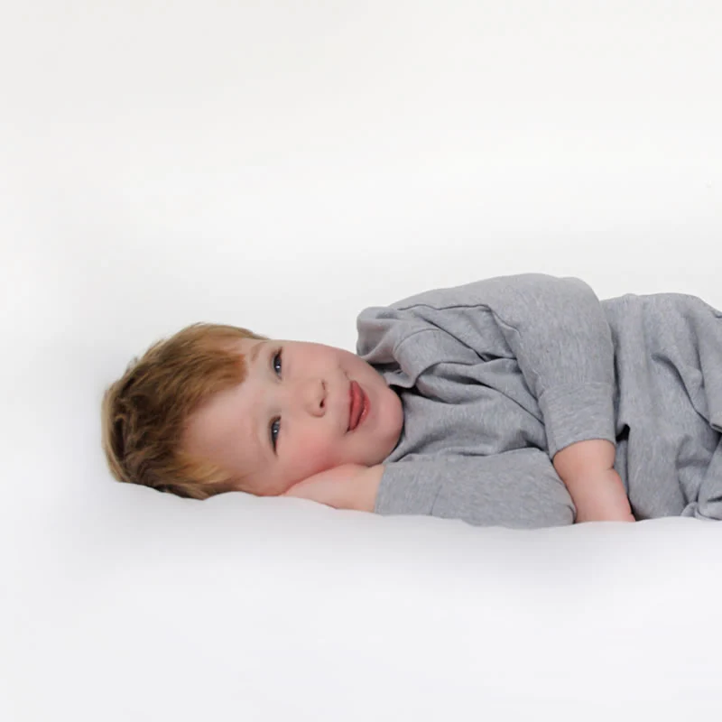 Young child with light brown hair lies on their side in a grey long-sleeved outfit, resting their head on one hand and looking content—perfect for a Baby Loves Sleep Hands In & Out Sleep Sack review—against a plain white background.