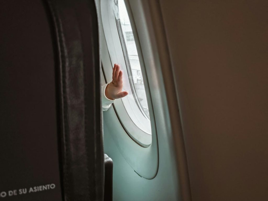 A childs hand is raised and pressed against the window of an airplane, seen from inside the cabin. The seatback and part of another seat are visible in the foreground.
