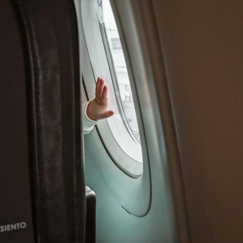 A childs hand is raised and pressed against the window of an airplane, seen from inside the cabin. The seatback and part of another seat are visible in the foreground.