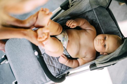 A baby wearing only a diaper lies in a gray stroller, looking up while an adult gently holds the babys feet. The scene is bright and softly lit.