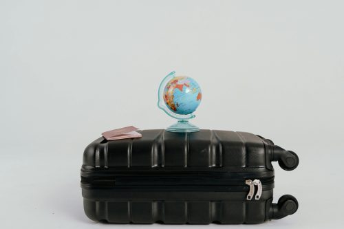 A small globe and a passport rest on top of a closed black suitcase with wheels, set against a plain white background.