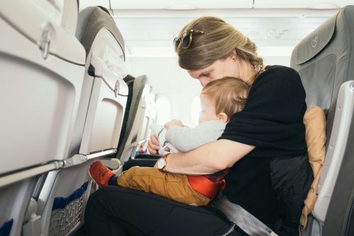 A woman sits in an airplane seat, embracing and comforting a small child on her lap. Both are wearing seat belts, and the woman has sunglasses on her head. The cabin interior and seats are visible around them.
