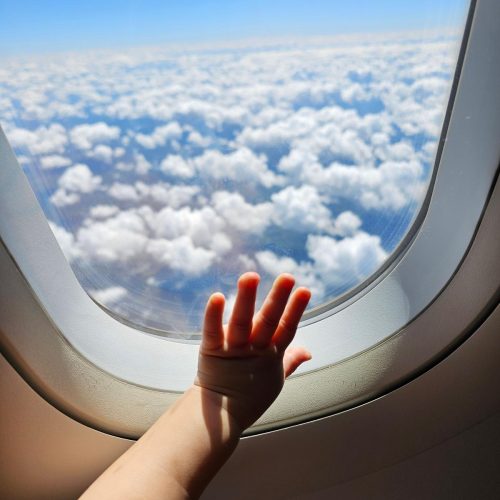 A childs hand reaches toward an airplane window, with fluffy white clouds and a clear blue sky visible outside.