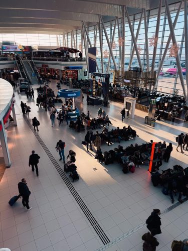 Crowds of people sit and walk in a bright, modern airport terminal with large windows, shops, seating areas, and a few planes visible outside. Sunlight streams in through the windows, casting long shadows.