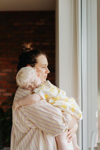 A woman in a striped shirt holds a sleeping toddler in a yellow dress, standing by a window with soft daylight. The scene conveys a sense of calm and care.