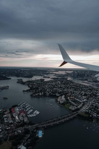 Airplane wing above a city with water, bridges, and scattered boats below, under a cloudy sky at dusk or dawn.